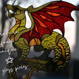 May include: A stained glass dragon suncatcher featuring textured yellow-green glass for the body and vibrant red-orange glass for the wings. The piece is constructed from 63 glass pieces using the Tiffany technique, with text reading Handmade in Tiffany Technique and 63 glass pieces.