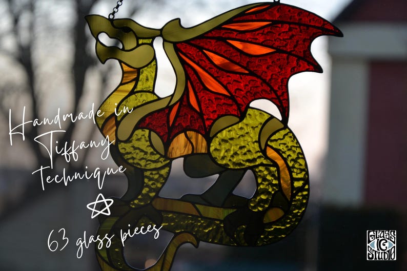 May include: A stained glass dragon suncatcher featuring textured yellow-green glass for the body and vibrant red-orange glass for the wings. The piece is constructed from 63 glass pieces using the Tiffany technique, with text reading Handmade in Tiffany Technique and 63 glass pieces.