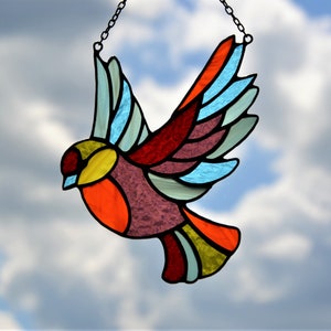 May include: A stained glass bird with a colorful design, hanging from a chain against a cloudy sky. The bird is made of red, orange, yellow, green, blue, and purple glass.