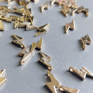 Op de afbeelding: Een strooiing van gouden bliksem-bedels op een grijs oppervlak. De bedels zijn klein en hebben een glanzende afwerking.