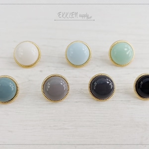 Puede incluir: Seis botones redondos de fondo plano con bordes dorados. Los botones son de varios colores: blanco, azul claro, verde claro, gris, negro y azul oscuro.