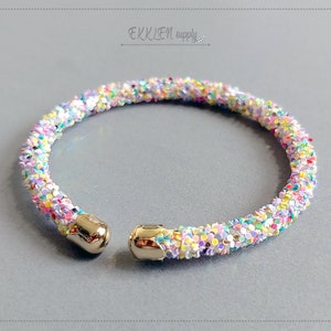 May include: A gold bangle bracelet with a colourful confetti design. The bracelet is made of small, iridescent beads that are arranged in a random pattern.