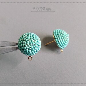 Puede incluir: Un par de pendientes de botón de cuentas de color azul turquesa con postes y bucles dorados. Las cuentas son pequeñas y están densamente empaquetadas, creando una superficie lisa y redondeada.