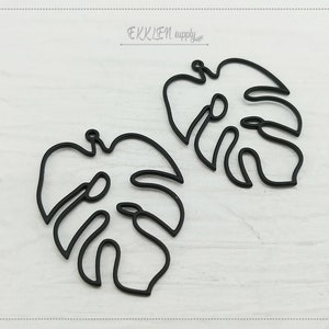 Könnte beinhalten: Zwei schwarze Metallanhänger in Form von Monstera-Blättern. Jeder Anhänger ist eine stilisierte Umrisslinie eines Blattes mit einem Loch zum Aufhängen. Die Anhänger befinden sich auf weißem Hintergrund. Der Text "EKKLEN supply" befindet sich oben links.