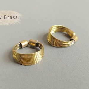 May include: Two gold-toned rings made of thin, braided wire. The rings are stacked on top of each other, with the top ring slightly offset to the right. The rings are on a gray surface. Raw Brass.