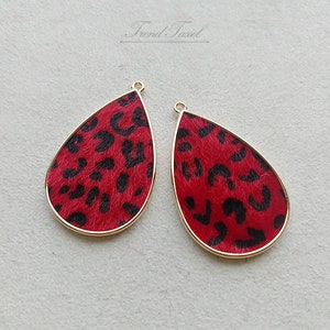 May include: Two teardrop-shaped earrings with a red leopard print design and gold-colored trim. The earrings have a small loop at the top for hanging. The background is a light gray color. The text "Trend Tasel" is in the upper right corner.