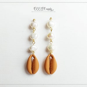 May include: A pair of dangle earrings with a brown cowrie shell charm and three white pearl beads on each earring.