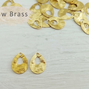 Może przedstawiać: Dwa złote metalowe charms w kształcie łzy z otworem na górze. Charms mają fakturę młotkową i leżą na białym drewnianym tle. W tle widoczny jest tekst "Raw Brass".