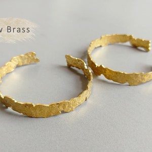 Puede incluir: Dos pulseras de latón en bruto de color dorado con una superficie texturizada irregular. Las pulseras están apiladas una encima de la otra.