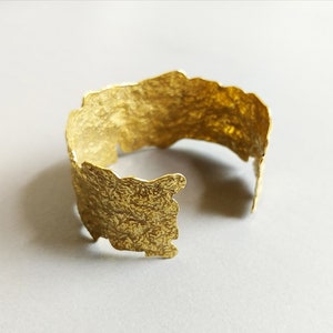 May include: A gold-toned metal bracelet with a textured, irregular surface. The bracelet is open and has a slightly curved shape.