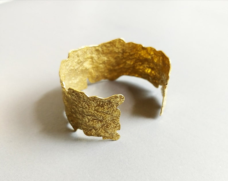 May include: A gold-toned metal bracelet with a textured, irregular surface. The bracelet is open and has a slightly curved shape.