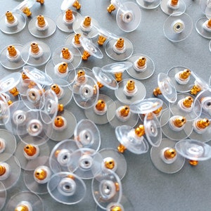 May include: A large group of clear plastic earring backs with gold-colored posts. The backs are scattered on a light gray surface.