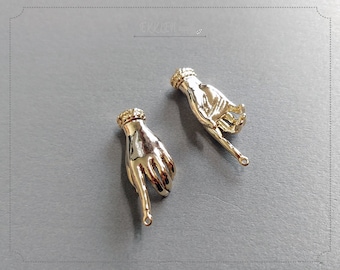 May include: Two gold-toned metal hand charms with a pointed finger pointing downwards. The hands have a textured surface and a gold-toned ring around the wrist.