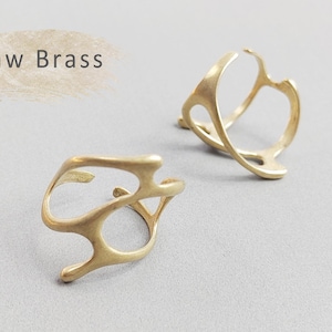 May include: Two raw brass rings with an organic, openwork design. The rings have an abstract, flowing shape, with the text "Raw Brass" in the upper left corner. The rings are displayed on a light gray surface.