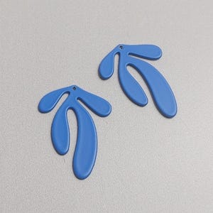 May include: Two blue leaf-shaped earrings. Each earring has three rounded lobes and a small hole at the top for attachment. The earrings are a solid, matte blue color and lie flat against a light gray surface.