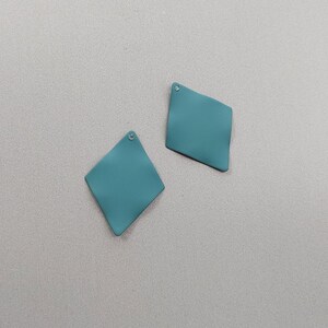 May include: Two teal diamond-shaped earring components. Each earring has a small hole at the top for attachment. The earrings have a slightly textured surface and a matte finish. The background is a light grey.