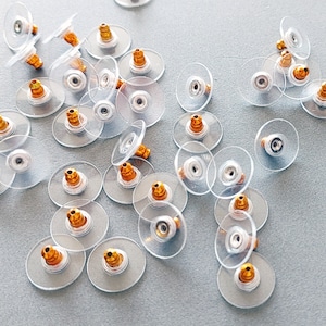 Puede incluir: Una colección de respaldos de pendientes de plástico transparente con postes de metal dorado. Los respaldos están dispuestos sobre una superficie gris claro.