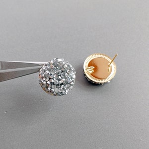 Op de afbeelding: Een paar zilverkleurige oorbellen met een rond, glinsterend, kristalontwerp. De oorbellen hebben een steek sluiting.