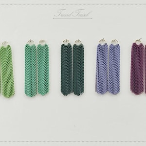 Puede incluir: Cinco pares de pendientes de borlas en varios colores: verde, turquesa, verde oscuro, morado y azul. Cada par presenta un aro plateado y un diseño de borla de cadena. El texto "Trend Tassel" es visible.