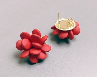 May include: Two red flower-shaped stud earrings. Each earring features a cluster of rounded petals in a vibrant red hue. The earring posts and backing are gold-colored, providing a contrasting accent. The earrings are set against a neutral gray background.