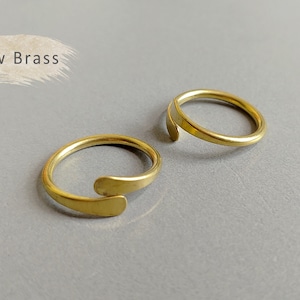 Puede incluir: Dos anillos abiertos y ajustables de latón en bruto. Los anillos tienen un diseño simple y minimalista con un acabado liso y pulido. Los anillos se muestran sobre una superficie gris. El texto "Raw Brass" está en la esquina superior izquierda.