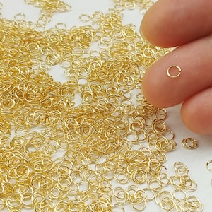 May include: A large quantity of small, gold-colored jump rings. The rings are scattered on a white surface. A hand is holding one of the rings to show its size.