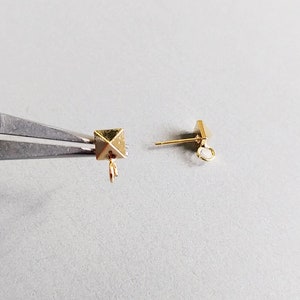 Pode incluir: Dois fechos de brinco dourados. Um fecho tem uma parte superior em forma de pirâmide e um laço na parte inferior. O outro fecho tem um poste reto com um laço na extremidade.