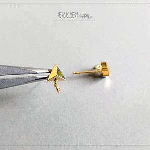 May include: Two gold triangle shaped earring findings with a loop on one end. The findings are shown on a gray surface.