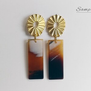 Puede incluir: Un par de pendientes de declaraci&oacute;n dorados y marrones. Los pendientes presentan un dise&ntilde;o de sol dorado con una abertura redonda en el centro y un colgante rectangular marr&oacute;n y blanco.