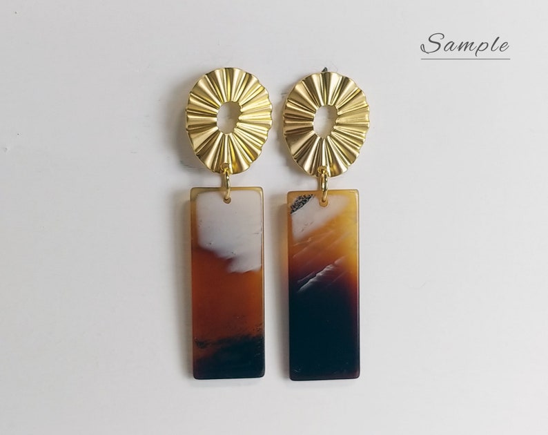 Puede incluir: Un par de pendientes de declaraci&oacute;n dorados y marrones. Los pendientes presentan un dise&ntilde;o de sol dorado con una abertura redonda en el centro y un colgante rectangular marr&oacute;n y blanco.