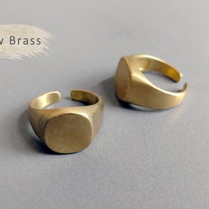 May include: Two simple, gold-toned, brass signet rings. The rings are flat and have a rounded, square shape. The text "Raw Brass" is visible in the top left corner of the image.