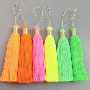 May include: Six colorful tassels in shades of orange, pink, yellow, and green. Each tassel has a gold-colored cap and a gold string loop for hanging. The tassels are made of a soft, fibrous material.