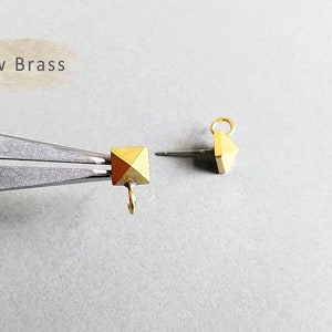 以下が含まれることがあります： ループが片側に、ピンが反対側に付いた、金色のピラミッド型の金属製チャームが2つ。画像には「Raw Brass」というテキストが表示されています。