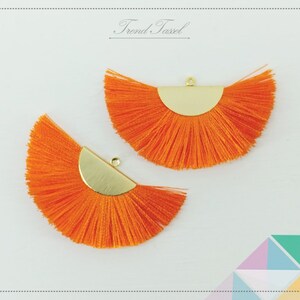 May include: Two orange tassel fan shapes with gold metal accents. The tassels are made of thin strands of yarn and are attached to a gold metal half-circle.