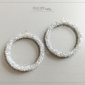 Può includere: Due orecchini circolari color argento. Ogni orecchino è incrostato di piccoli cristalli scintillanti, creando una superficie strutturata. Gli orecchini hanno un piccolo anello dorato nella parte superiore per appenderli. Lo sfondo è grigio chiaro e il testo "EKKLEN supply" è visibile.