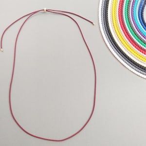 Pode incluir: Um colar de cordão bordô com detalhes em metal dourado. O colar é exibido em uma superfície cinza, com uma seleção de cordões coloridos em semicírculo ao fundo. Joias.