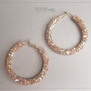 Könnte beinhalten: Ein Paar goldene Creolen mit glitzerndem Finish. Die Ohrringe sind groß und haben ein dickes, strukturiertes Design.