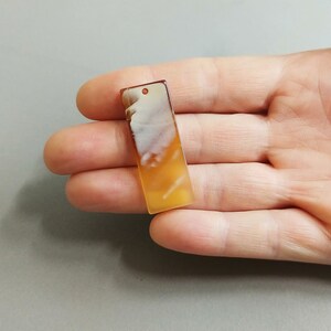 Puede incluir: Un colgante rectangular de &aacute;gata blanca y naranja con un agujero en la parte superior para encordar.