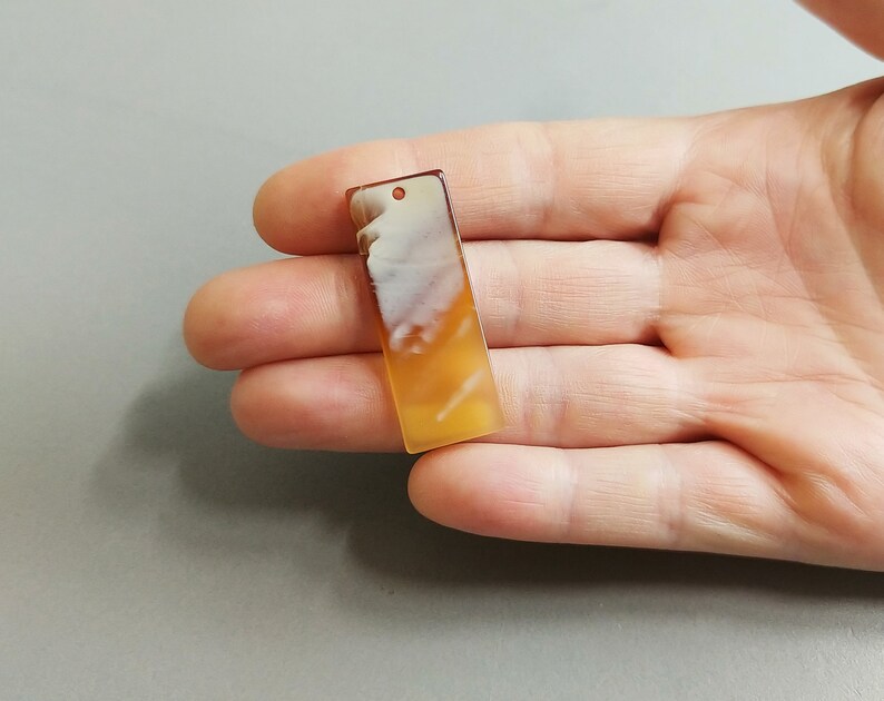Puede incluir: Un colgante rectangular de &aacute;gata blanca y naranja con un agujero en la parte superior para encordar.
