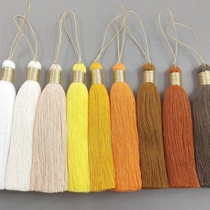 Könnte beinhalten: Acht farbenfrohe Quasten in Weiß-, Beige-, Gelb-, Orange- und Brauntönen. Jede Quaste hat ein goldfarbenes Metallband und eine goldene Schlaufe zum Aufhängen. Die Quasten bestehen aus weichem, strukturiertem Material.