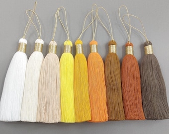 May include: A collection of eight colorful tassels in shades of white, beige, yellow, orange, and brown. Each tassel features a gold-colored metallic band at the top and a gold string loop for hanging. The tassels are made of soft, textured material.