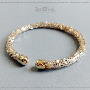 May include: A gold glitter bangle bracelet with a hinged clasp. The bracelet is made of small, round glitter pieces that are tightly packed together.