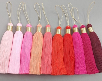 May include: A collection of colorful tassels in shades of pink, orange, and red. Each tassel has a gold-colored cap and a loop for hanging. The tassels are made of a soft, fringed material.