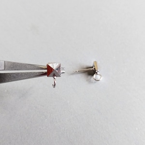 May include: Close-up of a silver earring with a pyramid-shaped charm and a small loop. The earring is being held in place by a pair of pliers.