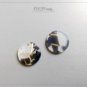 Könnte beinhalten: Zwei runde, weiße, schwarze und goldfarbene, gemusterte Harzembellen mit einem Loch zum Aufhängen.