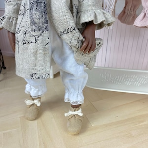 May include: A doll wearing a linen dress with a lace trim and black script print. The doll is wearing white pants with lace trim and brown shoes with white bows.