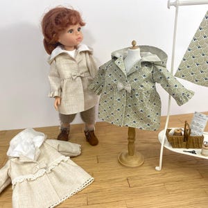 May include: Three doll coats in beige and green with white trim. The coats have different patterns and styles. The coats are displayed on a white stand with a sewing kit.