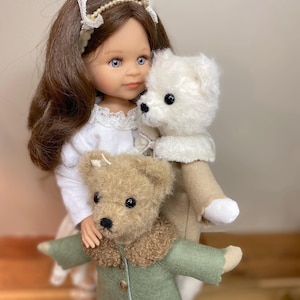 Puede incluir: Una muñeca con cabello castaño y ojos azules sostiene un oso de peluche blanco y un oso de peluche marrón que lleva un abrigo verde.