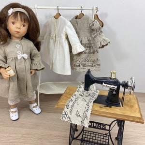 Puede incluir: Una muñeca con un abrigo de lino beige con un lazo blanco y sosteniendo un carrete de hilo. La muñeca está de pie frente a un estante blanco con dos vestidos de lino colgados. Una máquina de coser vintage está sobre una mesa de madera con una tela de lino en la parte superior.