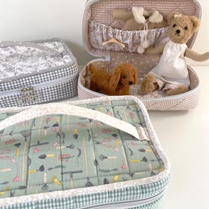 Peut inclure: Trois trousses décoratives en tissu avec des motifs à carreaux et floraux. Une trousse est ouverte, révélant un ours en peluche et une figurine de petit chien. La trousse ouverte a un motif vichy beige et blanc. Les deux autres trousses sont fermées, l'une avec un motif floral gris et l'autre avec un motif d'outils de jardinage verts.